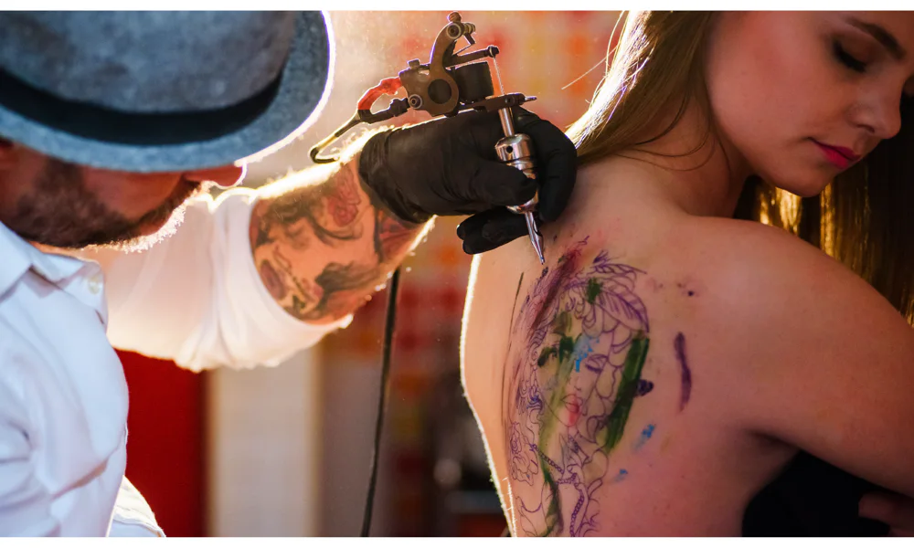 Tattoo artist applying a colorful watercolor tattoo design on a woman’s shoulder, with soft, fluid colors and no defined outlines.
