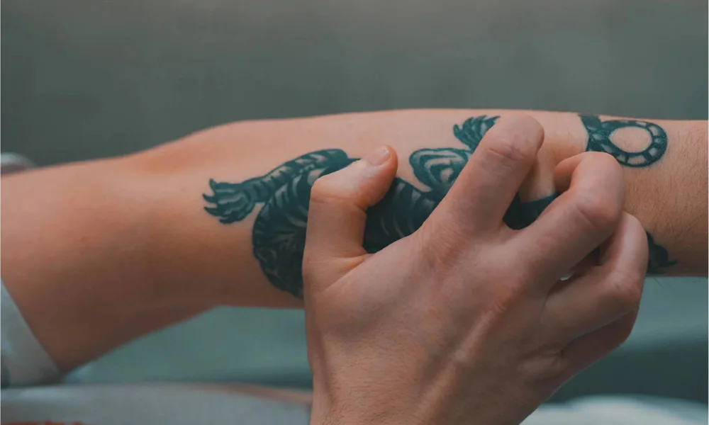 A close-up of a person scratching a tattoo on their arm, highlighting the importance of avoiding picking and scratching during the tattoo healing process to prevent damage and scarring.