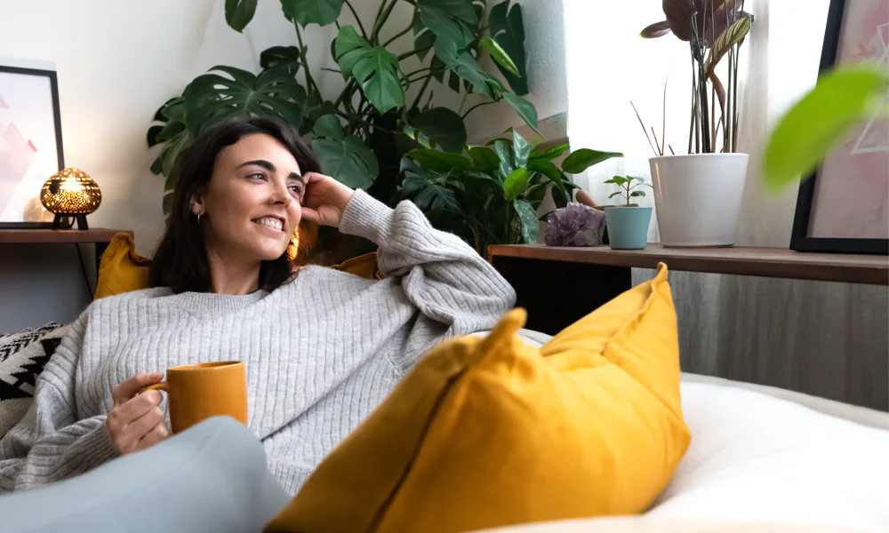 Relaxing scene of a woman enjoying a cup of tea on a couch, surrounded by plants, highlighting the importance of relaxation before getting a tattoo.