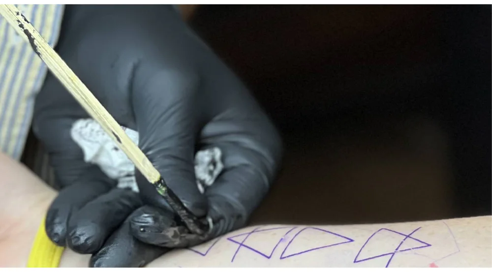 Close-up of hand-poked tattooing technique using a stick and ink, with a tattoo artist applying a simple geometric design on the skin.