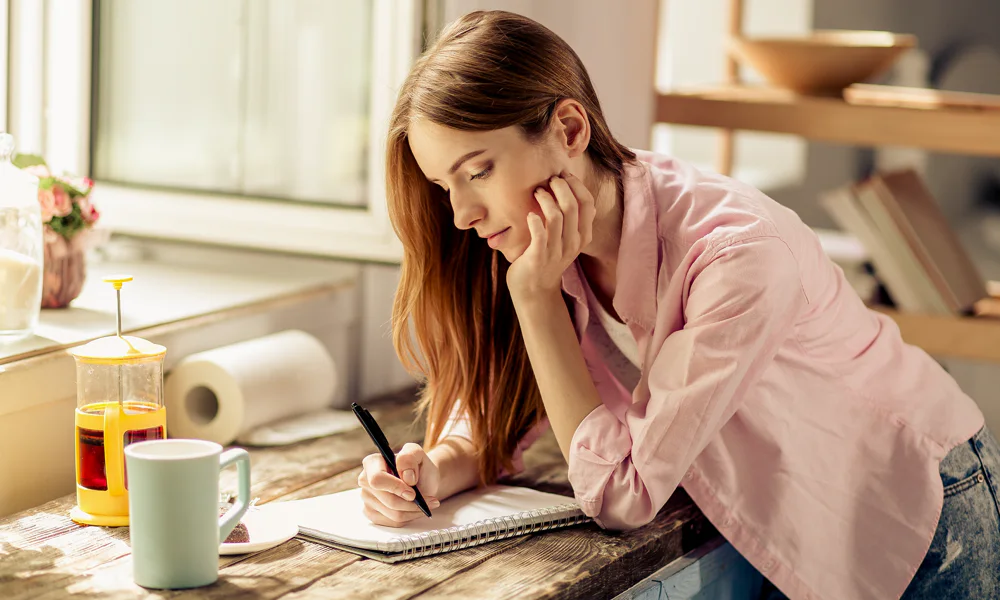A young woman writing notes in a cozy kitchen with natural light, while enjoying a drink next to her.