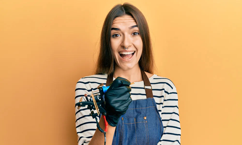 Choosing the Right Tattoo Artist - Smiling female tattoo artist holding a tattoo machine, wearing a striped shirt and apron, with an orange background.