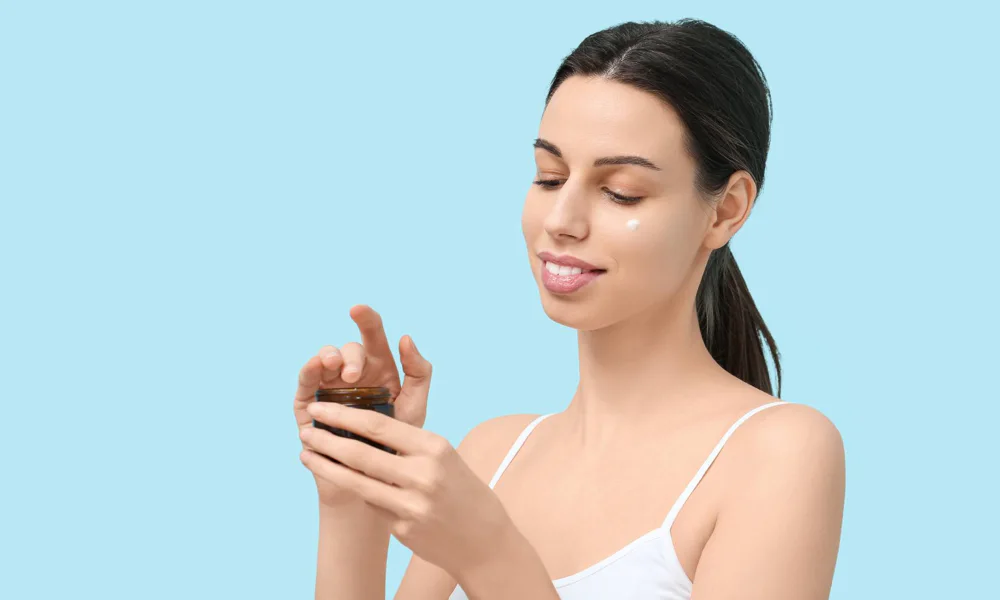 Skincare before getting a tattoo - Woman applying skincare cream, focusing on a small jar of product, against a light blue background.