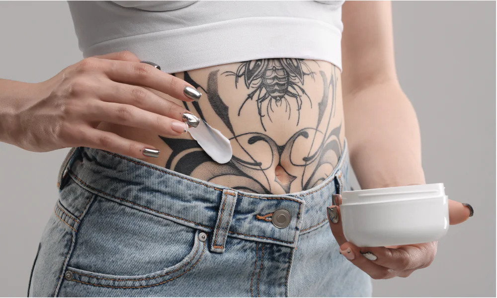 A person applying a moisturizing cream to a large tattoo on their stomach, showcasing proper tattoo aftercare to keep the ink vibrant and skin hydrated.