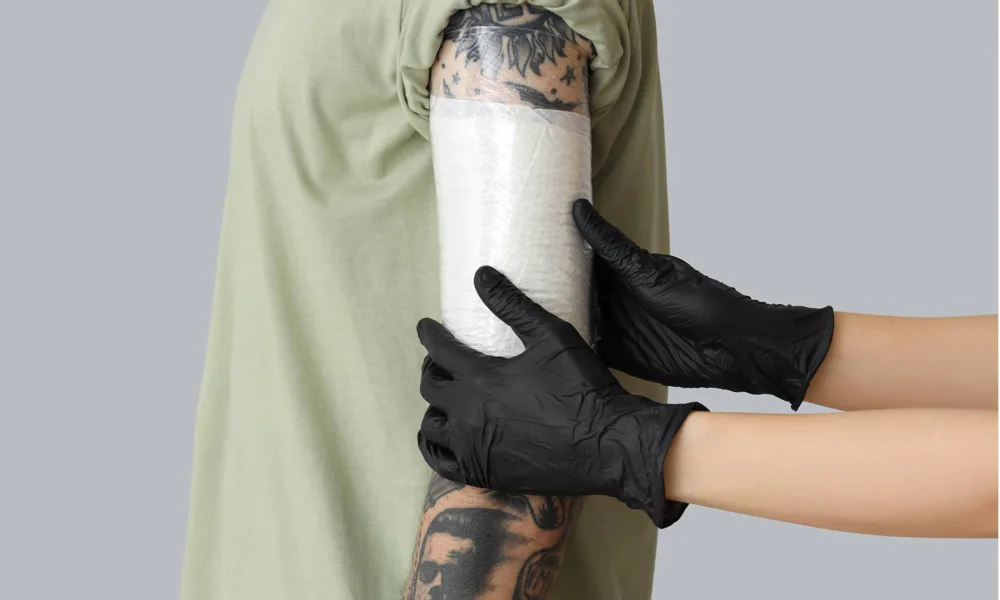 A tattoo artist, wearing black gloves, is applying a protective wrap to a freshly inked tattoo on a client's forearm. The tattoo, featuring intricate black-and-gray designs, is visible around the edges of the white wrap, ensuring the new ink stays clean and protected during the healing process.